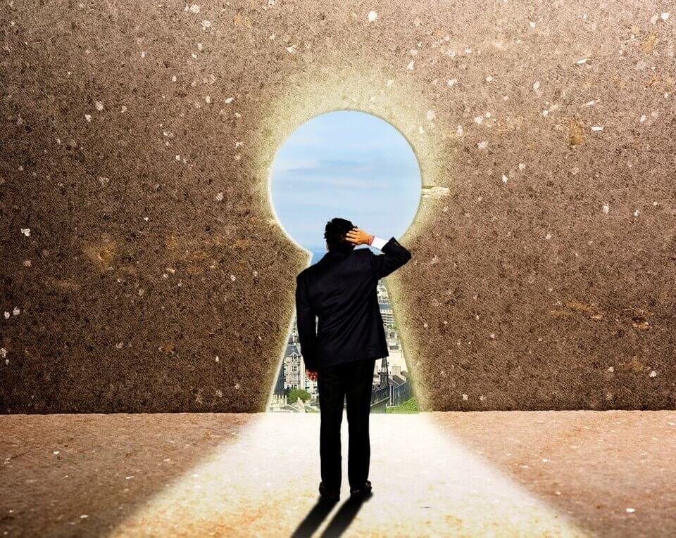 Mann von hinten im schwarzen Anzug, steht in einem Gebäude, rechte Hand am Hinterkopf fassend, der Raum befindet sich in der Höhe und hat eine Aussparung in Form eines Schlüsselloches, durch das der Mann schaut, Sonne scheint herein, er sieht blauen Himmel und andere Gebäude, er steht im hellen Lichtkegel